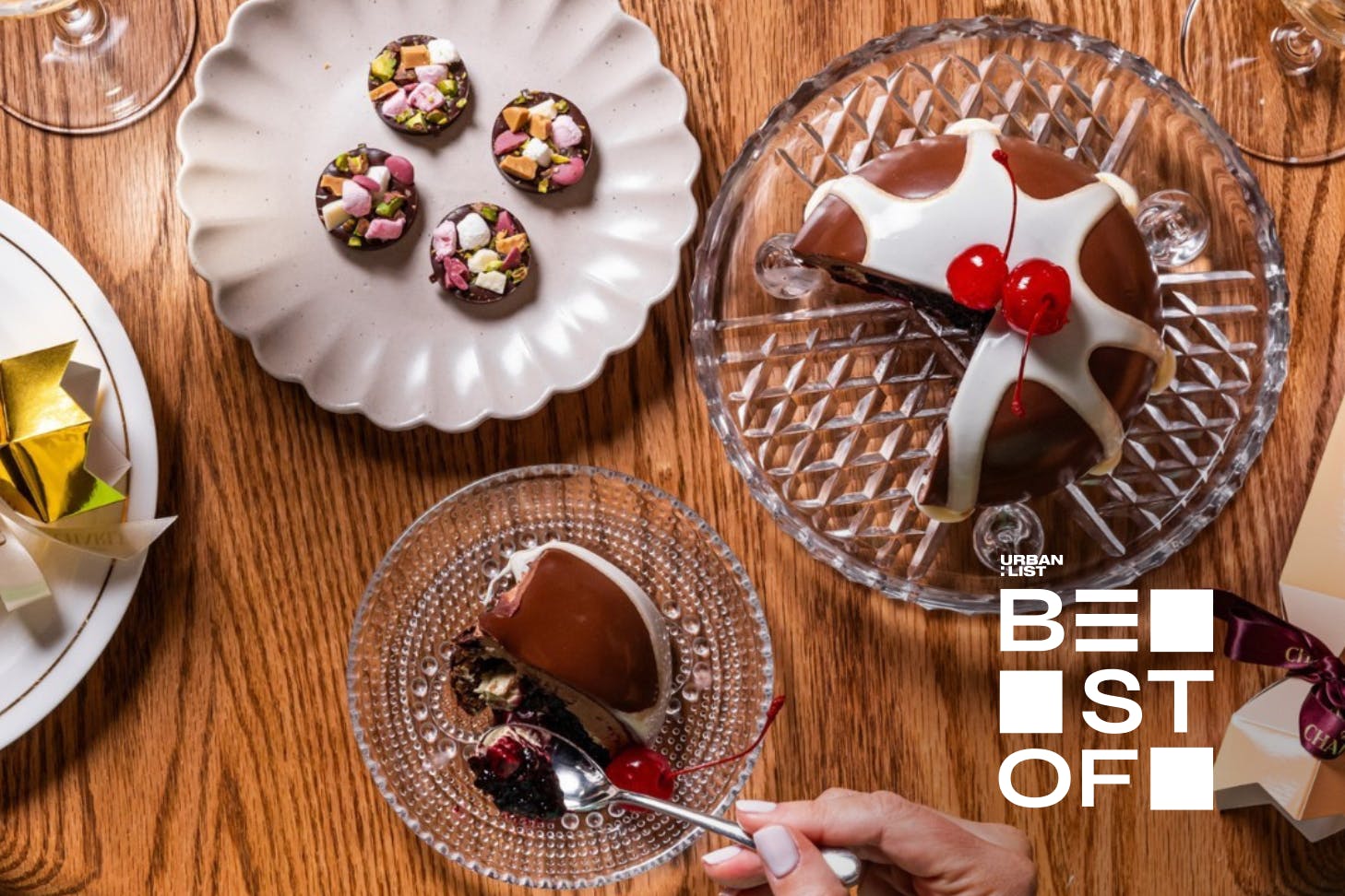 Overhead view of a festive dessert spread on a wooden table featuring a glossy chocolate Christmas pudding topped with cherries, bite-sized chocolates with nuts and marshmallows, and a hand holding a spoon ready to take a bite.